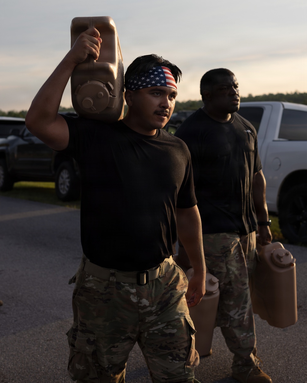 Two individuals in military-style attire are carrying tan/brown jerry cans in an outdoor parking area during low-light conditions, likely dawn or dusk. The foreground subject wears an American flag bandana, a black t-shirt, and camouflage pants while hoisting a jerry can overhead. The background subject similarly dressed carries a jerry can at his side near parked vehicles.