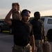 Two individuals in military-style attire are carrying tan/brown jerry cans in an outdoor parking area during low-light conditions, likely dawn or dusk. The foreground subject wears an American flag bandana, a black t-shirt, and camouflage pants while hoisting a jerry can overhead. The background subject similarly dressed carries a jerry can at his side near parked vehicles.