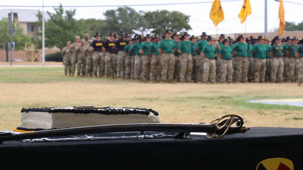 A military ceremony is taking place on an outdoor parade field, with a large formation of soldiers wearing green t-shirts, camouflage pants, and campaign/drill sergeant hats standing at attention in the background. In the foreground, a table covered with a black cloth displays a decorated white cake with black frosting trim and a cavalry saber (sword), suggesting a unit anniversary or birthday celebration. Yellow/gold guidon flags are visible in the background, consistent with U.S. Army cavalry unit traditions.