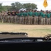 A military ceremony is taking place on an outdoor parade field, with a large formation of soldiers wearing green t-shirts, camouflage pants, and campaign/drill sergeant hats standing at attention in the background. In the foreground, a table covered with a black cloth displays a decorated white cake with black frosting trim and a cavalry saber (sword), suggesting a unit anniversary or birthday celebration. Yellow/gold guidon flags are visible in the background, consistent with U.S. Army cavalry unit traditions.