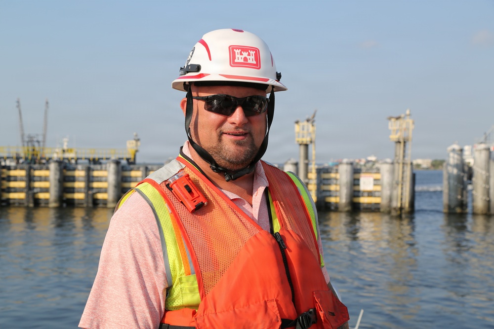 A male construction or engineering worker is photographed near a waterway infrastructure site. He is wearing standard safety gear including a white hard hat bearing the U.S. Army Corps of Engineers castle insignia, an orange high-visibility vest, and dark sunglasses. The background shows a marine construction or lock/dam structure with concrete pilings and yellow structural elements.