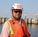 A male construction or engineering worker is photographed near a waterway infrastructure site. He is wearing standard safety gear including a white hard hat bearing the U.S. Army Corps of Engineers castle insignia, an orange high-visibility vest, and dark sunglasses. The background shows a marine construction or lock/dam structure with concrete pilings and yellow structural elements.