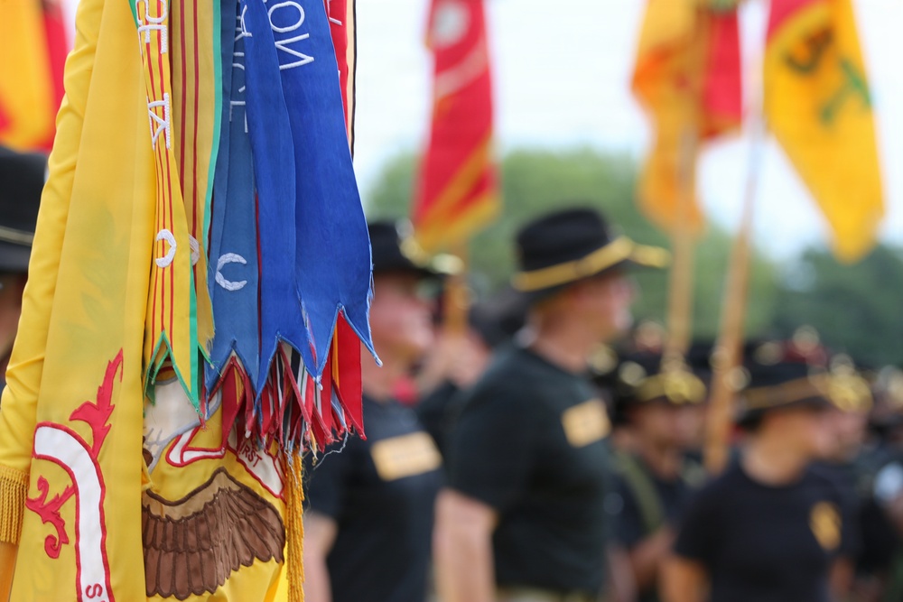 A military ceremony or parade featuring uniformed personnel carrying unit guidons and regimental flags. The foreground shows a yellow flag with red embroidery and an eagle emblem, adorned with multiple streamer ribbons in blue, red, and green colors. Personnel in dark uniforms and campaign hats are visible in the blurred background alongside additional colorful flags.