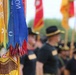 A military ceremony or parade featuring uniformed personnel carrying unit guidons and regimental flags. The foreground shows a yellow flag with red embroidery and an eagle emblem, adorned with multiple streamer ribbons in blue, red, and green colors. Personnel in dark uniforms and campaign hats are visible in the blurred background alongside additional colorful flags.