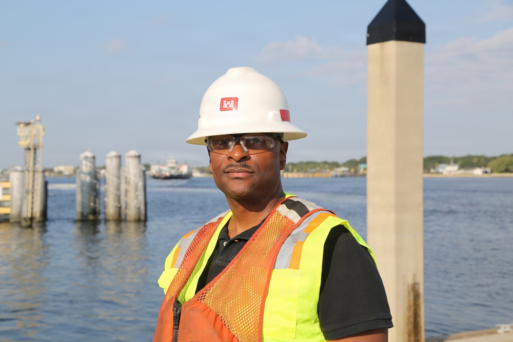 A construction or safety worker is posed near a waterfront or dock area wearing standard personal protective equipment. The background shows a body of water with dock pilings and a distant shoreline. The image appears to be a professional or promotional portrait taken in natural daylight.