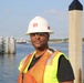 A construction or safety worker is posed near a waterfront or dock area wearing standard personal protective equipment. The background shows a body of water with dock pilings and a distant shoreline. The image appears to be a professional or promotional portrait taken in natural daylight.