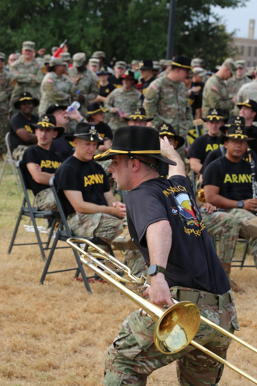 A U.S. Army musician in the foreground holds a trombone and adjusts a black cavalry hat with gold trim during an outdoor military event. Behind him, rows of soldiers are seated in folding chairs wearing black 'ARMY' t-shirts and matching black cavalry hats, while additional uniformed soldiers in camouflage stand in the background. The setting appears to be a military installation ceremony or gathering.