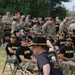 A U.S. Army musician in the foreground holds a trombone and adjusts a black cavalry hat with gold trim during an outdoor military event. Behind him, rows of soldiers are seated in folding chairs wearing black 'ARMY' t-shirts and matching black cavalry hats, while additional uniformed soldiers in camouflage stand in the background. The setting appears to be a military installation ceremony or gathering.