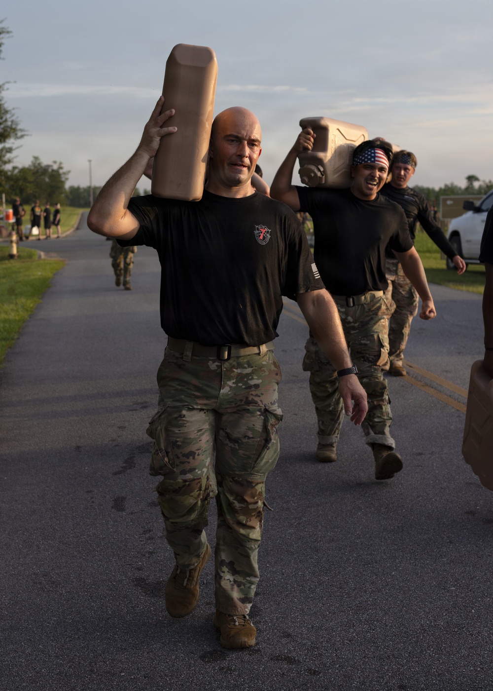 A group of military personnel in camouflage uniforms and black t-shirts are conducting a physical training exercise, carrying what appear to be weighted objects or jerry cans overhead while moving along a paved road. The lead individual, a bald man wearing a black t-shirt with a Special Forces insignia, carries a tan-colored object above his head with a strained expression. The setting appears to be a military installation during early morning or evening hours based on the lighting.