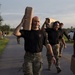 A group of military personnel in camouflage uniforms and black t-shirts are conducting a physical training exercise, carrying what appear to be weighted objects or jerry cans overhead while moving along a paved road. The lead individual, a bald man wearing a black t-shirt with a Special Forces insignia, carries a tan-colored object above his head with a strained expression. The setting appears to be a military installation during early morning or evening hours based on the lighting.