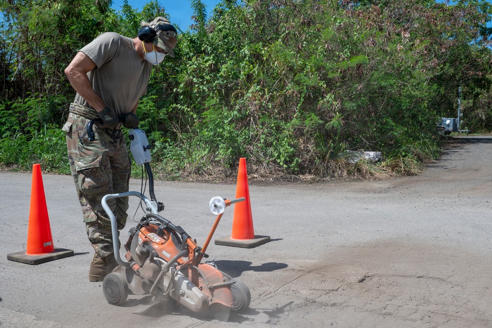 A person in military camouflage uniform is operating an orange concrete/pavement cutting saw on a road surface. The work area is marked with orange traffic safety cones, and the operator is wearing appropriate personal protective equipment including a respirator mask, ear protection, and gloves. Dust is visible rising from the cutting blade as it works through the pavement.