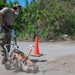 A person in military camouflage uniform is operating an orange concrete/pavement cutting saw on a road surface. The work area is marked with orange traffic safety cones, and the operator is wearing appropriate personal protective equipment including a respirator mask, ear protection, and gloves. Dust is visible rising from the cutting blade as it works through the pavement.