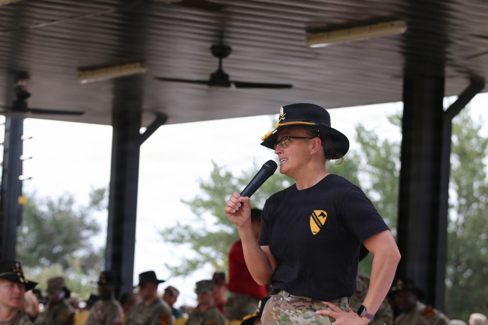 A military officer wearing a black campaign hat and 1st Cavalry Division t-shirt speaks into a microphone at an outdoor covered pavilion. The speaker appears to be addressing a large formation of soldiers visible in the background. The setting suggests a formal military ceremony or command address event.