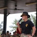 A military officer wearing a black campaign hat and 1st Cavalry Division t-shirt speaks into a microphone at an outdoor covered pavilion. The speaker appears to be addressing a large formation of soldiers visible in the background. The setting suggests a formal military ceremony or command address event.