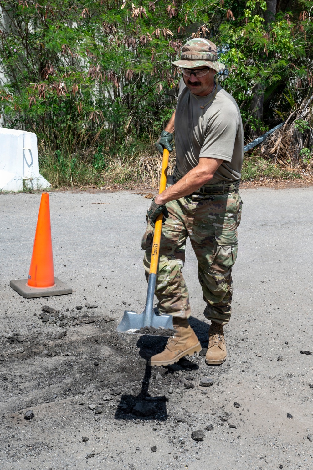 A military service member in OCP (Operational Camouflage Pattern) uniform is performing road repair work, using a square-blade shovel to work asphalt into a pothole on a paved surface. The individual is wearing safety glasses, work gloves, a boonie hat, and tan combat boots. An orange traffic cone is visible nearby, indicating a work zone.