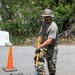 A military service member in OCP (Operational Camouflage Pattern) uniform is performing road repair work, using a square-blade shovel to work asphalt into a pothole on a paved surface. The individual is wearing safety glasses, work gloves, a boonie hat, and tan combat boots. An orange traffic cone is visible nearby, indicating a work zone.