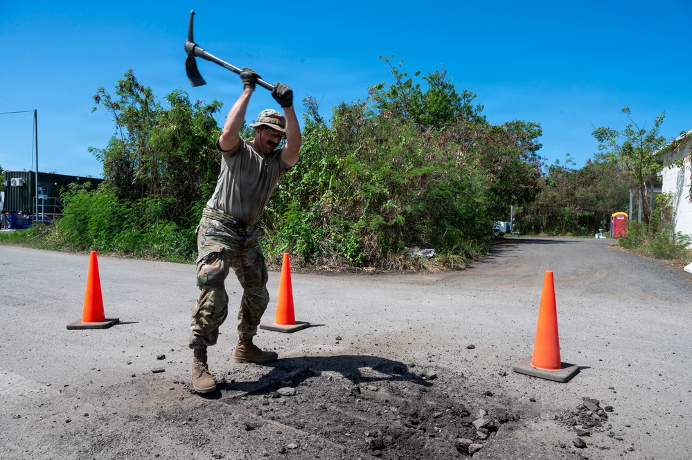 A person in military-style camouflage clothing and a boonie hat is using a pickaxe to break up damaged asphalt on a road. Three orange traffic cones are positioned around the work area to mark the hazard zone. The setting appears to be a tropical or subtropical location, with lush green vegetation visible in the background.