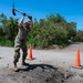 A person in military-style camouflage clothing and a boonie hat is using a pickaxe to break up damaged asphalt on a road. Three orange traffic cones are positioned around the work area to mark the hazard zone. The setting appears to be a tropical or subtropical location, with lush green vegetation visible in the background.