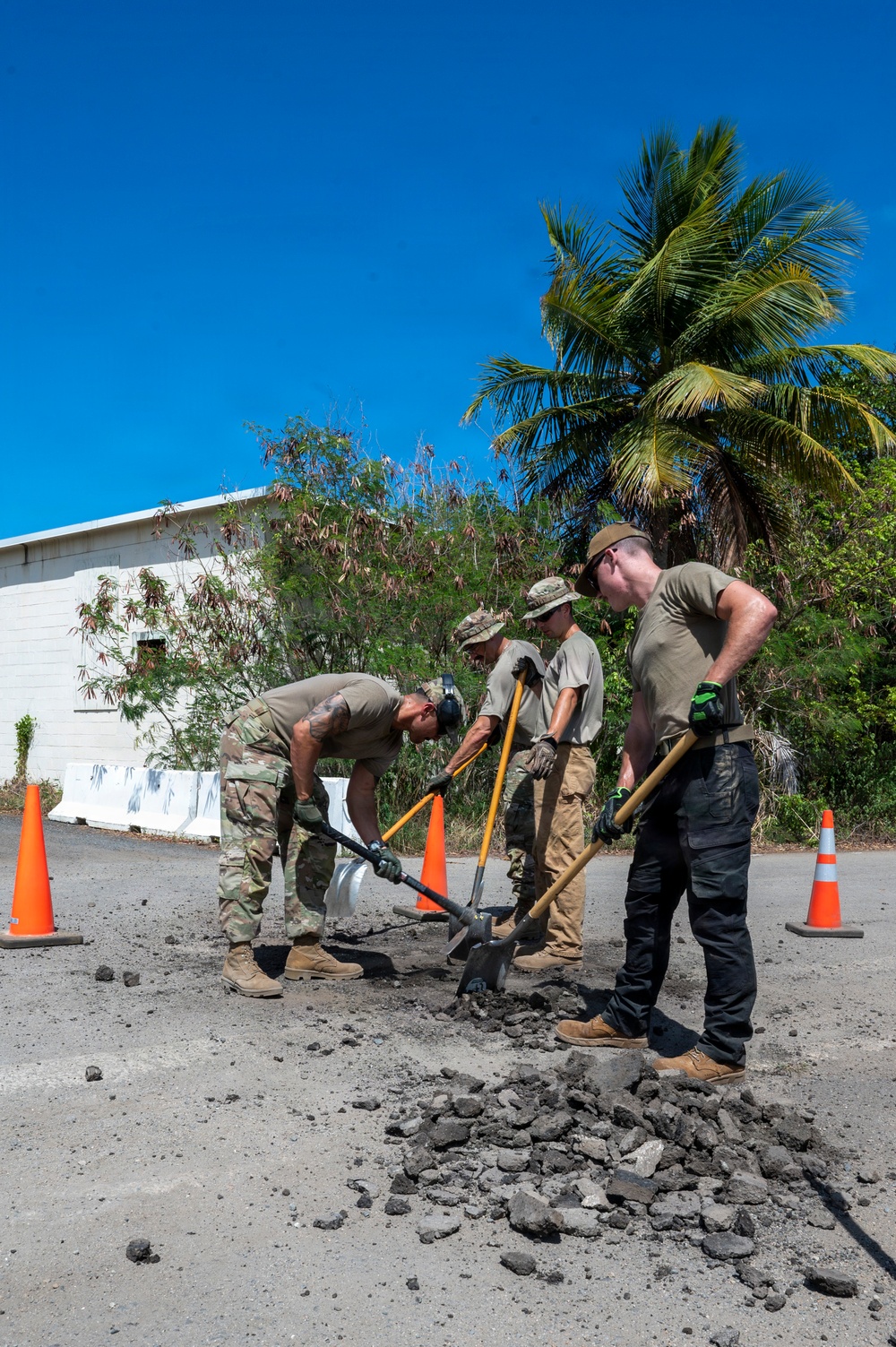 Four military personnel in uniform are performing road repair work, breaking up and removing damaged asphalt from a paved surface. The work site is marked with orange traffic cones and is located near a white building and tropical vegetation, suggesting a warm-climate military installation. The team is using shovels and a pickaxe to excavate a pothole or damaged road section.