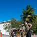 Four military personnel in uniform are performing road repair work, breaking up and removing damaged asphalt from a paved surface. The work site is marked with orange traffic cones and is located near a white building and tropical vegetation, suggesting a warm-climate military installation. The team is using shovels and a pickaxe to excavate a pothole or damaged road section.