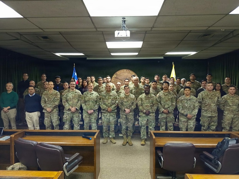 A large group of military personnel and a few civilians pose for a formal group photograph in what appears to be a conference or briefing room. Behind them is a seal or emblem on the wall reading 'INSTITUTO GEOGRAFICO' along with two flags, one of which appears to be the Chilean flag. The setting suggests a joint military or geographic institute event, likely involving both Chilean and U.S. military personnel.