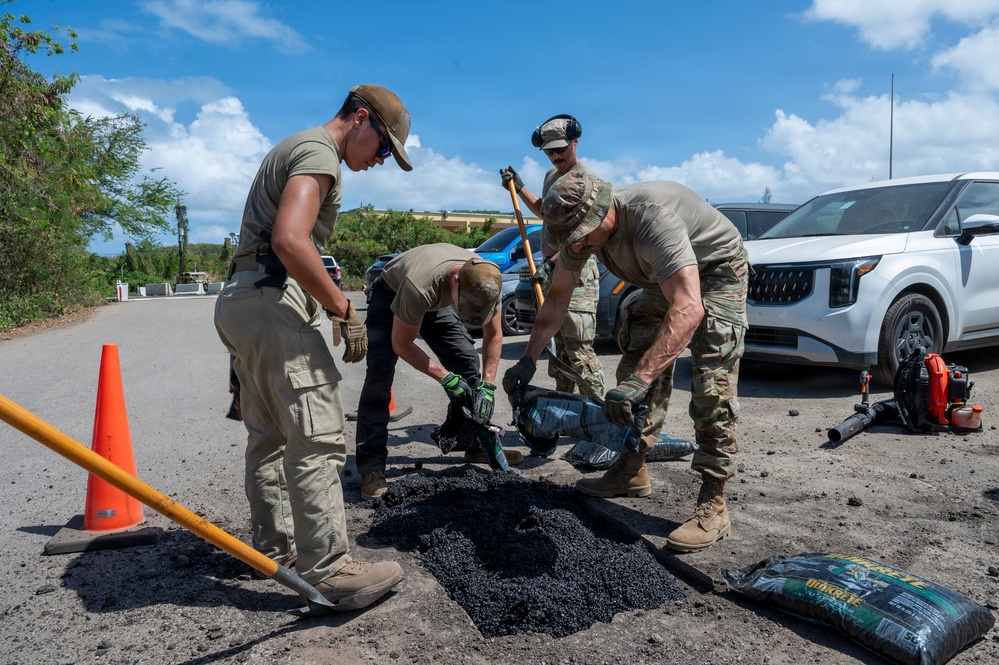 A group of four individuals in military-style uniforms are repairing a pothole in a road using bagged asphalt patching material. They are working collaboratively, using shovels and hand tools to fill and compact the damaged road surface. The scene appears to be a road maintenance or disaster recovery operation conducted in a tropical or subtropical environment.