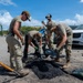 A group of four individuals in military-style uniforms are repairing a pothole in a road using bagged asphalt patching material. They are working collaboratively, using shovels and hand tools to fill and compact the damaged road surface. The scene appears to be a road maintenance or disaster recovery operation conducted in a tropical or subtropical environment.
