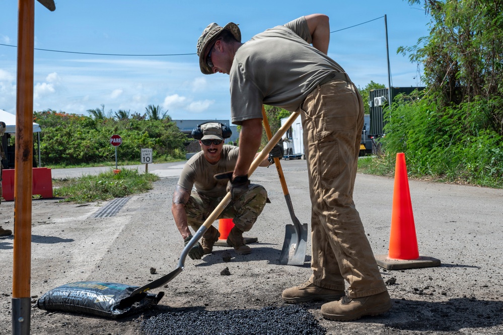 Two military personnel in tan/multicam uniforms are performing road repair work, filling a pothole with asphalt material. One individual is crouching and spreading material while the other stands and uses a shovel to work the asphalt. The work site is marked with orange traffic cones and is located near a road with visible signage in the background.