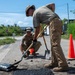 Two military personnel in tan/multicam uniforms are performing road repair work, filling a pothole with asphalt material. One individual is crouching and spreading material while the other stands and uses a shovel to work the asphalt. The work site is marked with orange traffic cones and is located near a road with visible signage in the background.