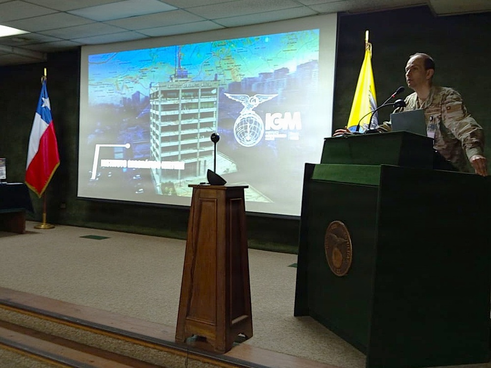 A military officer in camouflage uniform is presenting at a podium in a formal conference room. The presentation slide behind him displays the 'IGM' (Instituto Geográfico Militar) logo along with an aerial image of a building and a map background. The Chilean flag is visible on the left side of the room, indicating this event is taking place in Chile.