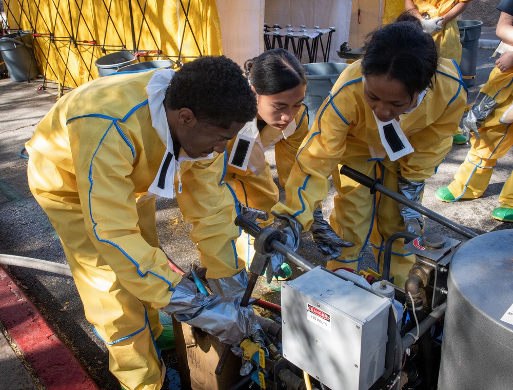 Three individuals wearing yellow protective hazmat-style suits with blue trim and black gloves are bent over operating or inspecting mechanical equipment outdoors on a paved surface. A visible 'DANGER 120 VOLTS' warning label is affixed to a metal electrical box on the equipment. The setting appears to be a training exercise or drill, indicated by the organized setup with yellow tents, gray barrels, and additional similarly-suited personnel in the background.