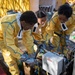Three individuals wearing yellow protective hazmat-style suits with blue trim and black gloves are bent over operating or inspecting mechanical equipment outdoors on a paved surface. A visible 'DANGER 120 VOLTS' warning label is affixed to a metal electrical box on the equipment. The setting appears to be a training exercise or drill, indicated by the organized setup with yellow tents, gray barrels, and additional similarly-suited personnel in the background.
