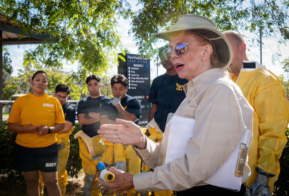A woman in a tan shirt and wide-brimmed hat appears to be briefing or instructing a group of U.S. Navy personnel outside the Naval Medical Center San Diego. She is holding a clipboard and a hose nozzle, suggesting a training or emergency preparedness exercise. Several personnel are wearing yellow protective gear, indicating a hazmat or disaster response drill.