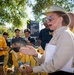 A woman in a tan shirt and wide-brimmed hat appears to be briefing or instructing a group of U.S. Navy personnel outside the Naval Medical Center San Diego. She is holding a clipboard and a hose nozzle, suggesting a training or emergency preparedness exercise. Several personnel are wearing yellow protective gear, indicating a hazmat or disaster response drill.