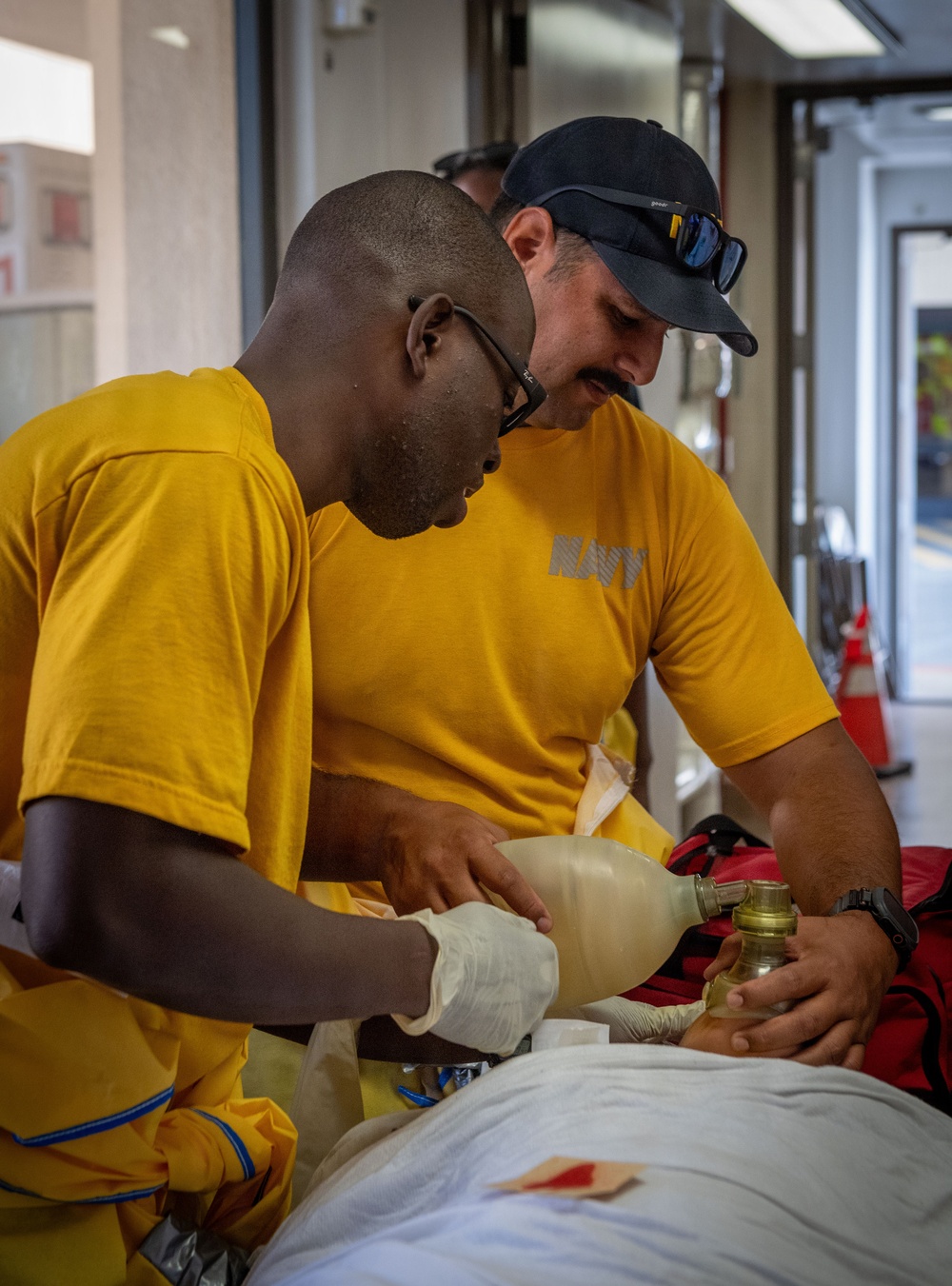 Two U.S. Navy personnel in yellow 'NAVY' PT shirts are performing what appears to be a medical training exercise. They are administering bag-valve-mask (BVM) ventilation on a mannequin or simulated patient lying on a surface. A red medical bag is visible nearby, suggesting a first aid or emergency response drill.