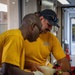 Two U.S. Navy personnel in yellow 'NAVY' PT shirts are performing what appears to be a medical training exercise. They are administering bag-valve-mask (BVM) ventilation on a mannequin or simulated patient lying on a surface. A red medical bag is visible nearby, suggesting a first aid or emergency response drill.