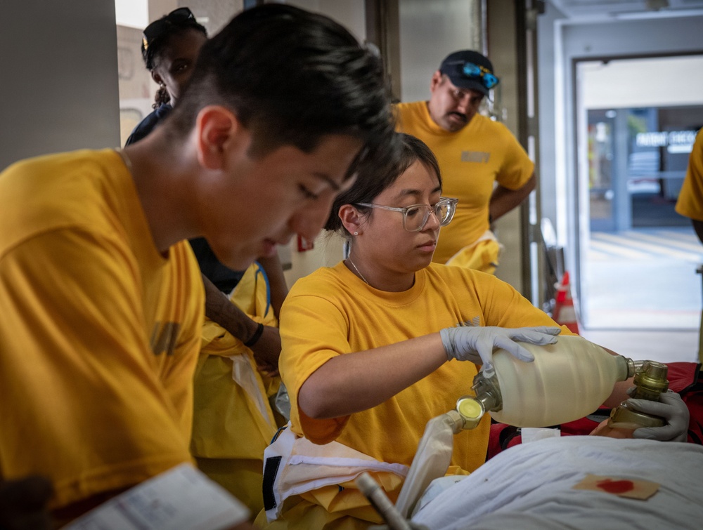 A group of individuals wearing matching yellow t-shirts are engaged in what appears to be a training or operational exercise involving equipment handling. A woman wearing clear-framed glasses and white latex gloves is manipulating what appears to be a gas cylinder or pressurized vessel with brass fittings. The setting appears to be near a building entrance or loading area.