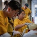 A group of individuals wearing matching yellow t-shirts are engaged in what appears to be a training or operational exercise involving equipment handling. A woman wearing clear-framed glasses and white latex gloves is manipulating what appears to be a gas cylinder or pressurized vessel with brass fittings. The setting appears to be near a building entrance or loading area.