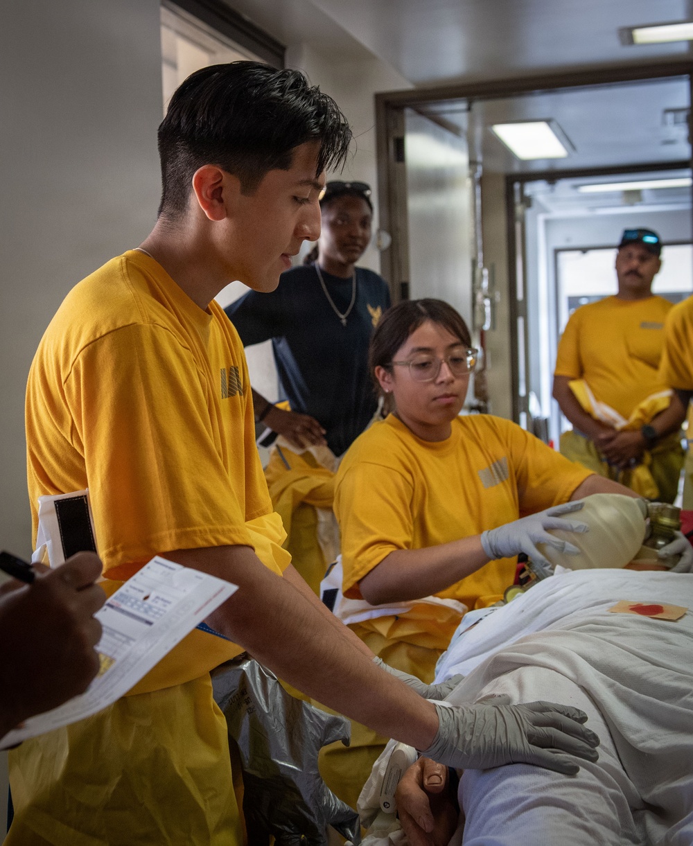 A group of individuals wearing matching yellow t-shirts appear to be conducting a medical training exercise or drill, attending to a patient on a stretcher or gurney. At least two people in yellow shirts are wearing latex gloves and actively working on the patient, while others observe. The setting appears to be an indoor corridor or medical facility, possibly aboard a naval vessel or institutional building.