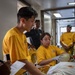 A group of individuals wearing matching yellow t-shirts appear to be conducting a medical training exercise or drill, attending to a patient on a stretcher or gurney. At least two people in yellow shirts are wearing latex gloves and actively working on the patient, while others observe. The setting appears to be an indoor corridor or medical facility, possibly aboard a naval vessel or institutional building.