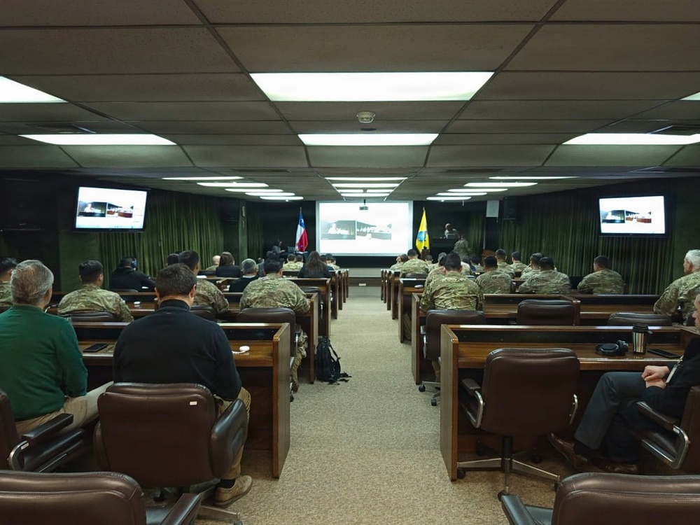 A military briefing or conference is taking place in a formal auditorium-style room. The majority of attendees are wearing military camouflage uniforms, seated at rows of wooden desks facing a projection screen at the front. Two flags are visible near the presenter's area, and supplementary monitors display the same presentation content on both sides of the room.