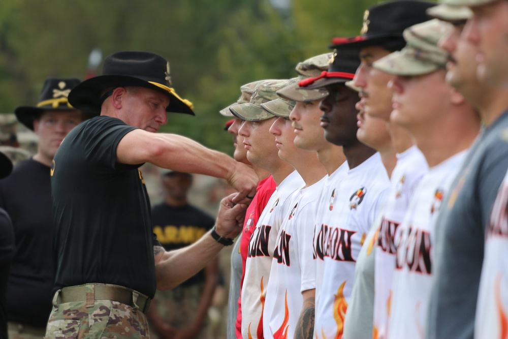 A military officer wearing a black cavalry hat and multicam uniform is shaking hands or presenting something to a line of individuals wearing white baseball jerseys with flame designs and military caps. The setting appears to be an outdoor military ceremony or recognition event. Personnel in Army uniforms are visible in the background observing the proceedings.