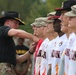 A military officer wearing a black cavalry hat and multicam uniform is shaking hands or presenting something to a line of individuals wearing white baseball jerseys with flame designs and military caps. The setting appears to be an outdoor military ceremony or recognition event. Personnel in Army uniforms are visible in the background observing the proceedings.