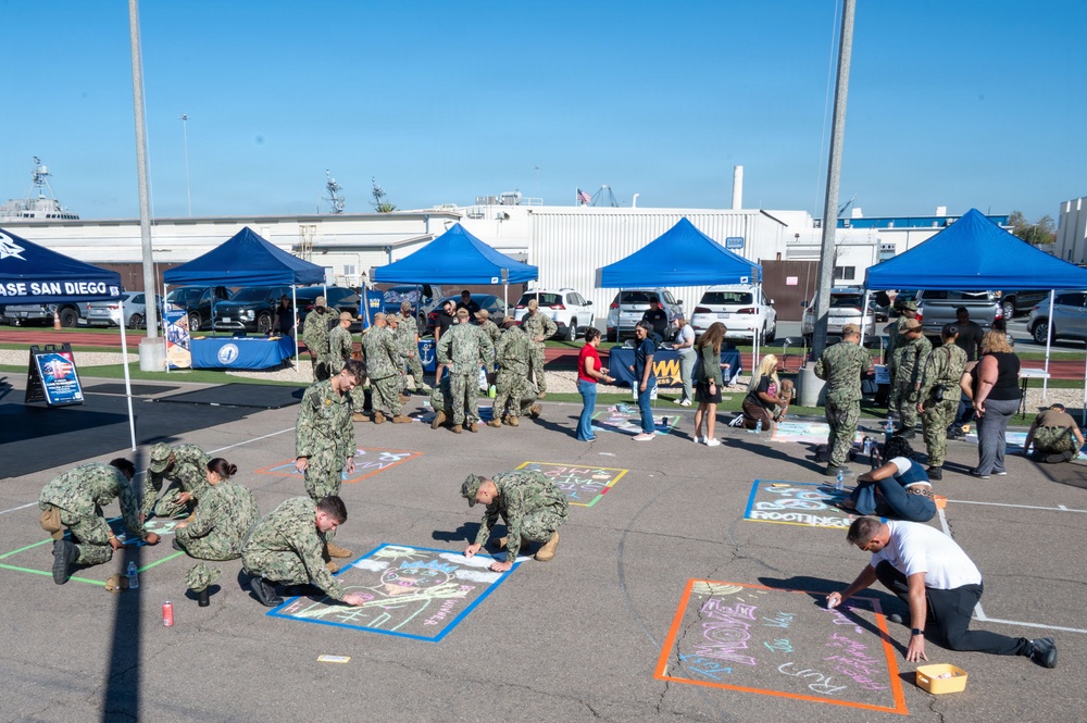 A chalk art event is taking place at Naval Base San Diego, with military personnel in camouflage uniforms kneeling on a paved surface creating chalk drawings within taped-off rectangular sections. Multiple blue canopy tents are set up in the background, suggesting an organized outdoor community or morale event. Both uniformed service members and civilians are participating or observing the activity.