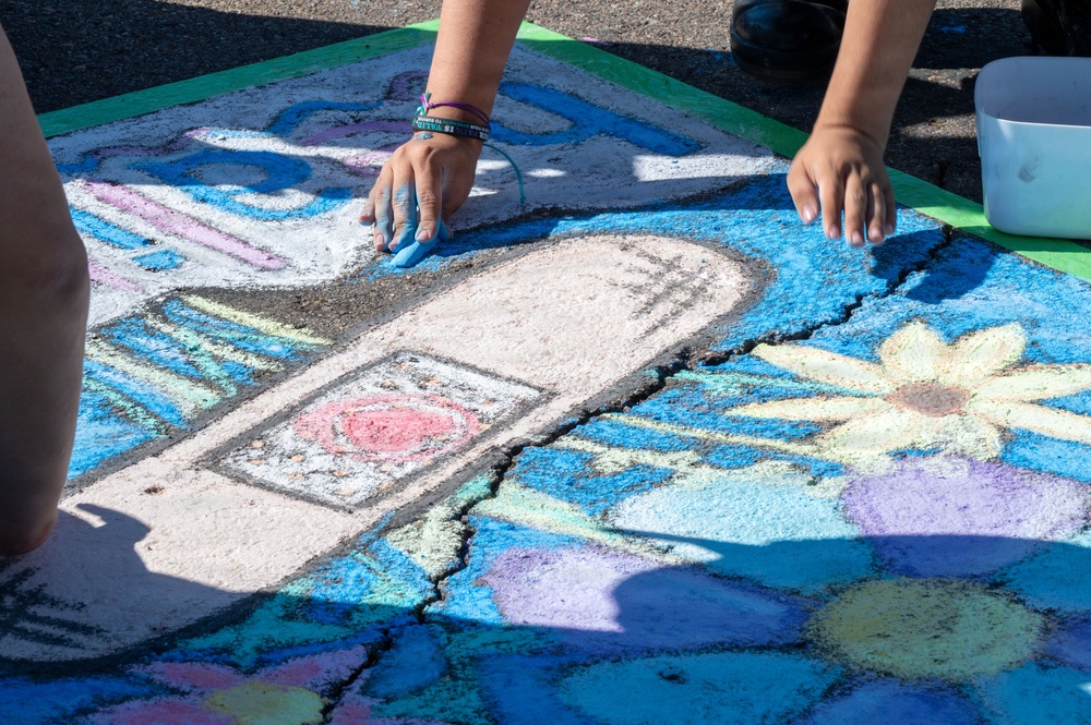 Two individuals are creating a colorful chalk art mural on a paved surface, likely during a street art festival or outdoor event. The artwork features a prominent white elongated shape resembling a menstrual pad or bandage, surrounded by flowers, blue backgrounds, and various colorful designs. The artists' chalk-stained hands are actively blending and applying chalk to the pavement.