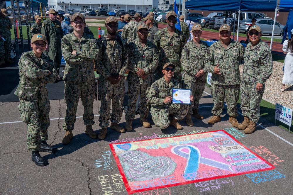 A group of U.S. Navy personnel in NWU (Naval Working Uniform) camouflage are posing together outdoors at what appears to be a military awareness event. One sailor is kneeling in front holding a certificate of achievement, and behind them is a colorful chalk art drawing on the pavement related to suicide prevention awareness. A sign reading 'Don't Give Up Fighting' is visible in the background near a blue tent.