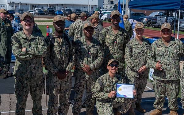 A group of U.S. Navy personnel in NWU (Naval Working Uniform) camouflage are posing together outdoors at what appears to be a military awareness event. One sailor is kneeling in front holding a certificate of achievement, and behind them is a colorful chalk art drawing on the pavement related to suicide prevention awareness. A sign reading 'Don't Give Up Fighting' is visible in the background near a blue tent.