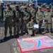 A group of U.S. Navy personnel in NWU (Naval Working Uniform) camouflage are posing together outdoors at what appears to be a military awareness event. One sailor is kneeling in front holding a certificate of achievement, and behind them is a colorful chalk art drawing on the pavement related to suicide prevention awareness. A sign reading 'Don't Give Up Fighting' is visible in the background near a blue tent.