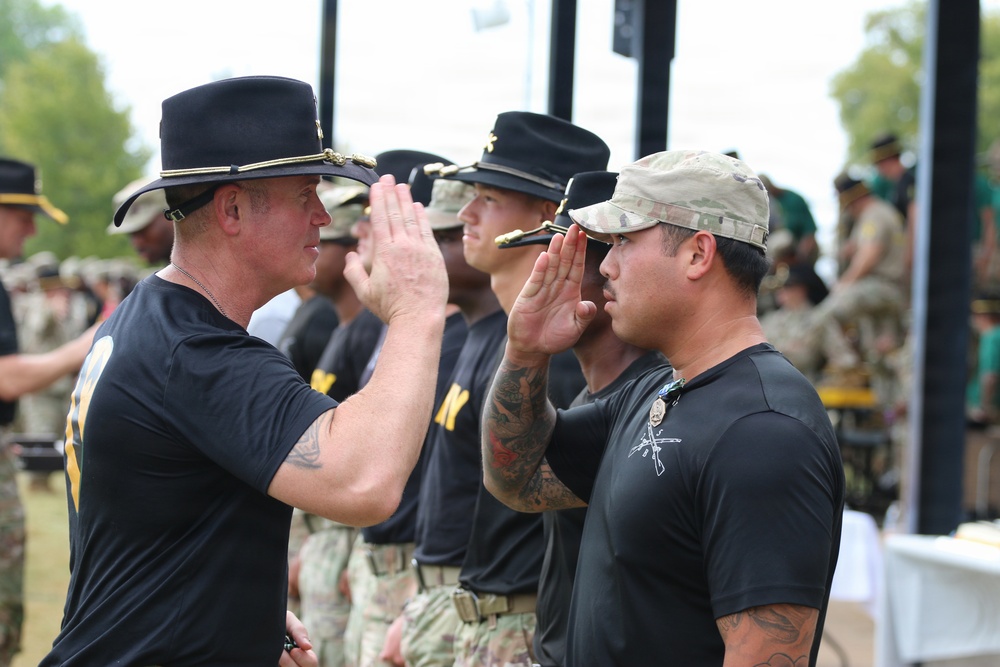 A military ceremony is taking place outdoors where personnel in black PT (physical training) shirts and military camouflage pants are raising their right hands, likely taking an oath or being sworn in. A senior figure wearing a black cavalry-style Stetson hat is administering the ceremony to a line of soldiers. The event appears to be a formal military reenlistment or induction ceremony conducted in an open-air setting.