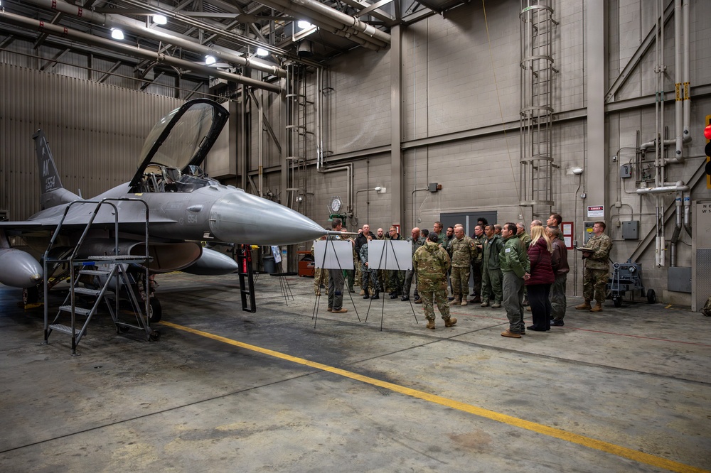 A group of approximately 20-25 military and civilian personnel are gathered inside a large aircraft maintenance hangar for a briefing or presentation. They are standing in front of two easel-mounted whiteboards/flip charts near an F-16 Fighting Falcon fighter jet with tail markings 'AK' and number '554'. The setting appears to be a U.S. Air Force facility, likely in Alaska based on the 'AK' tail code.